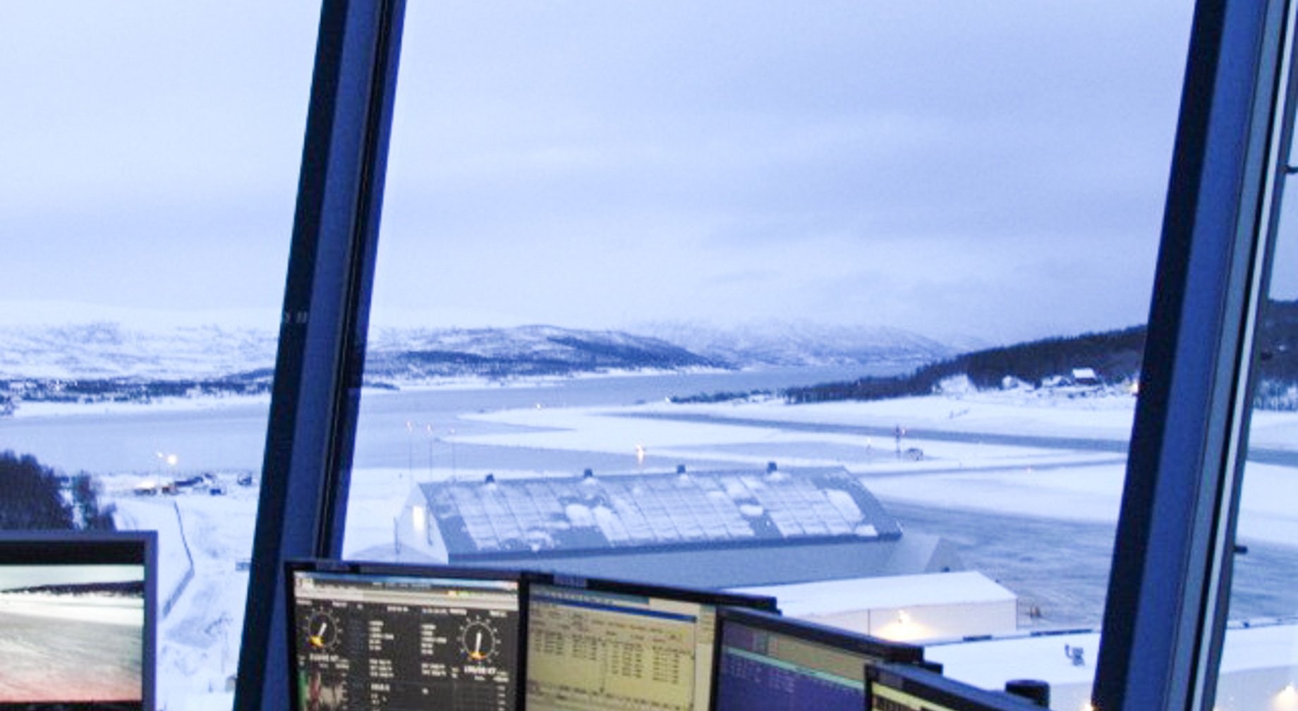 Utsikt fra flygekontrolltårn mot snødekt flyplass og fjellandskap i Norge, med flere dataskjermer i forgrunnen.