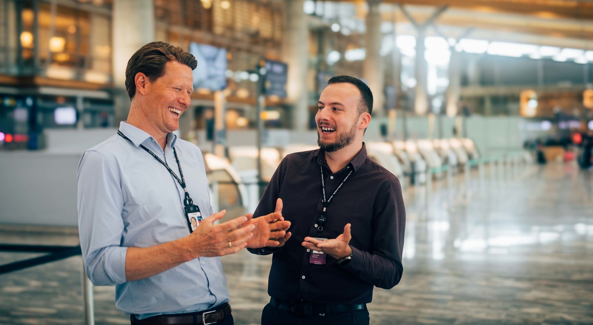 Two men laughing and talking together at a modern airport. They have ID marking around their necks, which indicates a professional connection. Bright and open surroundings with check-in areas in the background.