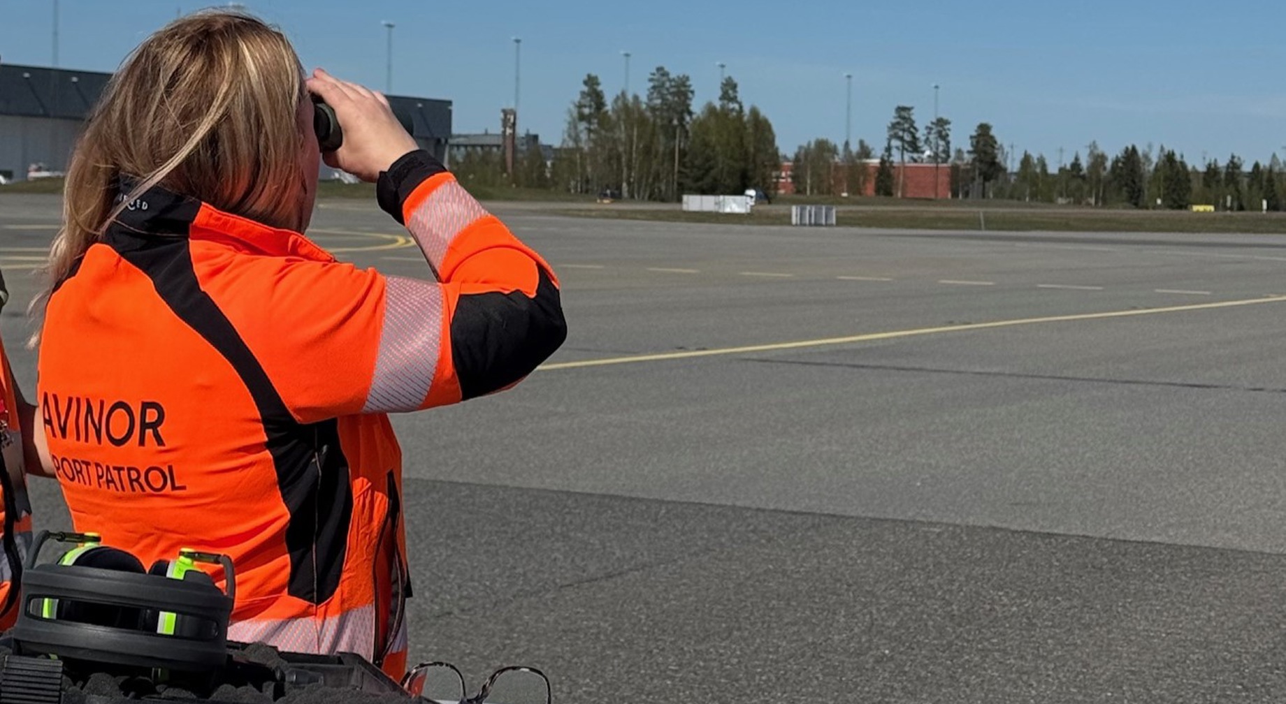 Avinor flyplasspatrulje i oransje refleksklær observerer rullebanen med kikkert på en flyplass.