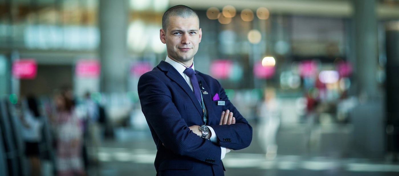 Mann i dress i en moderne flyplassterminal, med armene i kors og et selvsikkert uttrykk, med fokus på service og profesjonalitet.