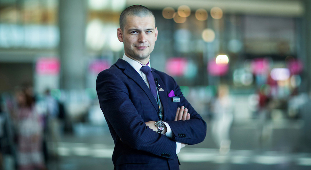 Mann i dress i en moderne flyplassterminal, med armene i kors og et selvsikkert uttrykk, med fokus på service og profesjonalitet.