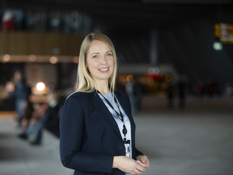 Kvinne med blondt hår i formell dressjakke smiler i en moderne konferansesenter. Blury bakgrunn med mennesker og belysning.