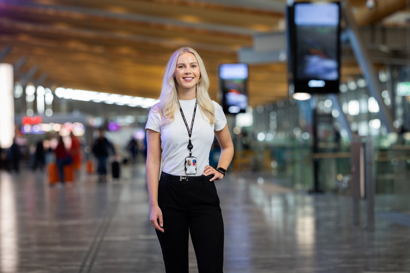 Kvinne i sort bukse og hvit t-skjorte på flyplass; står i moderne terminal, smilende med besøkskort rundt halsen.