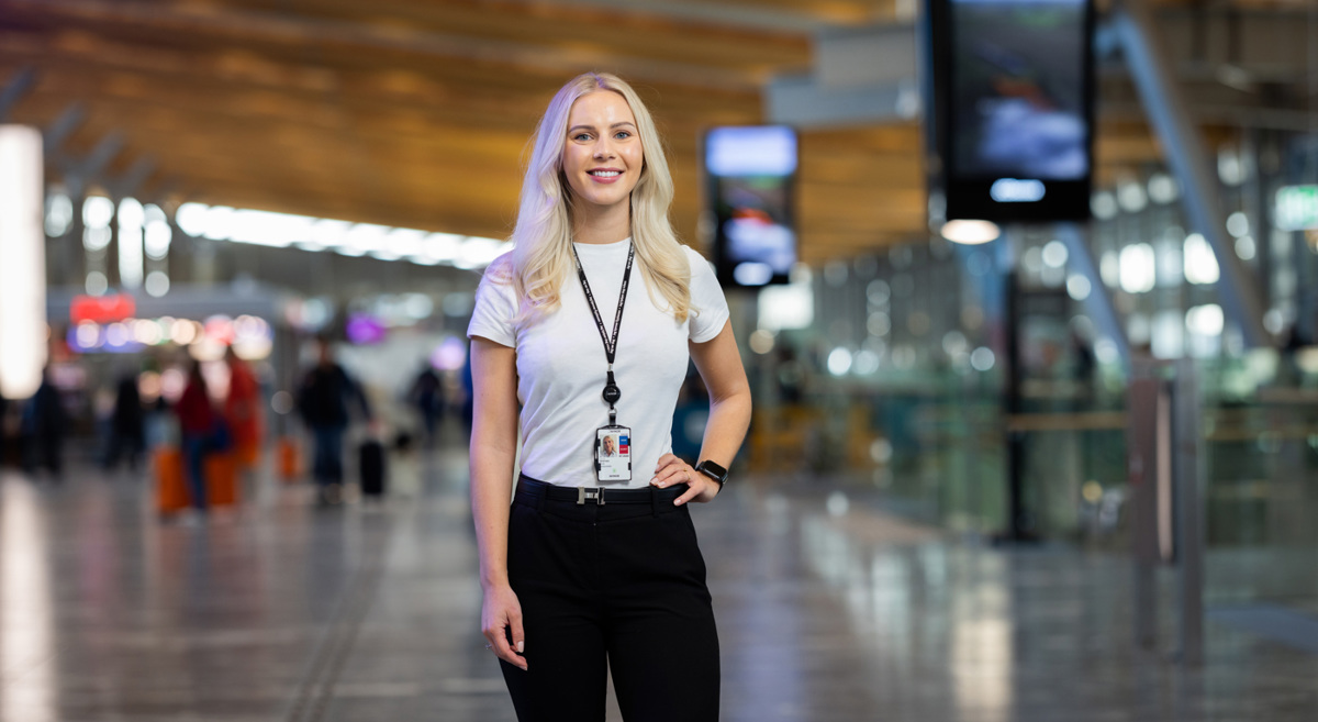 Kvinne i sort bukse og hvit t-skjorte på flyplass; står i moderne terminal, smilende med besøkskort rundt halsen.