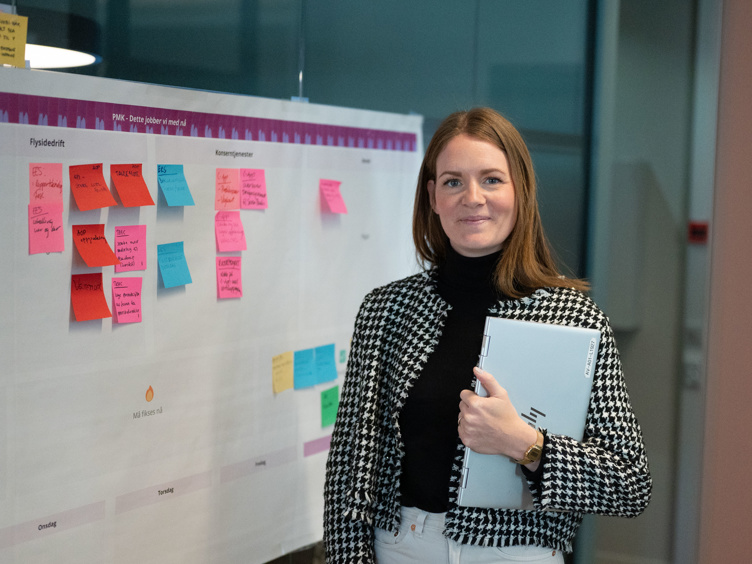 Kvinne som står foran en tavle med fargerike post-it-lapper, holder en datamaskin under armen, ikledd svart og hvit rutete jakke