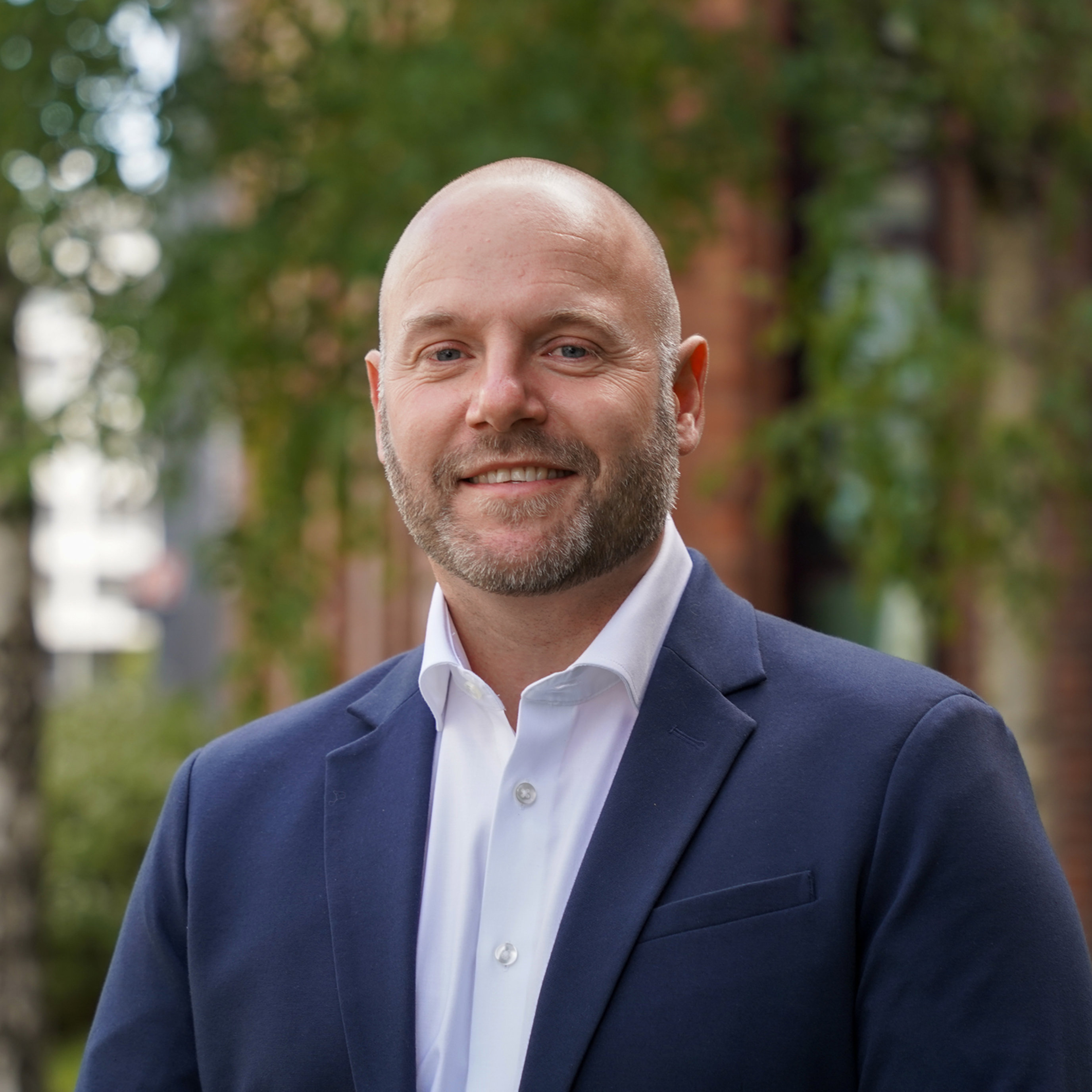 Person i formell blå dressjakke og kvit skjorte, smilande utandørs framfor grøne tre og ei raud murbygning.