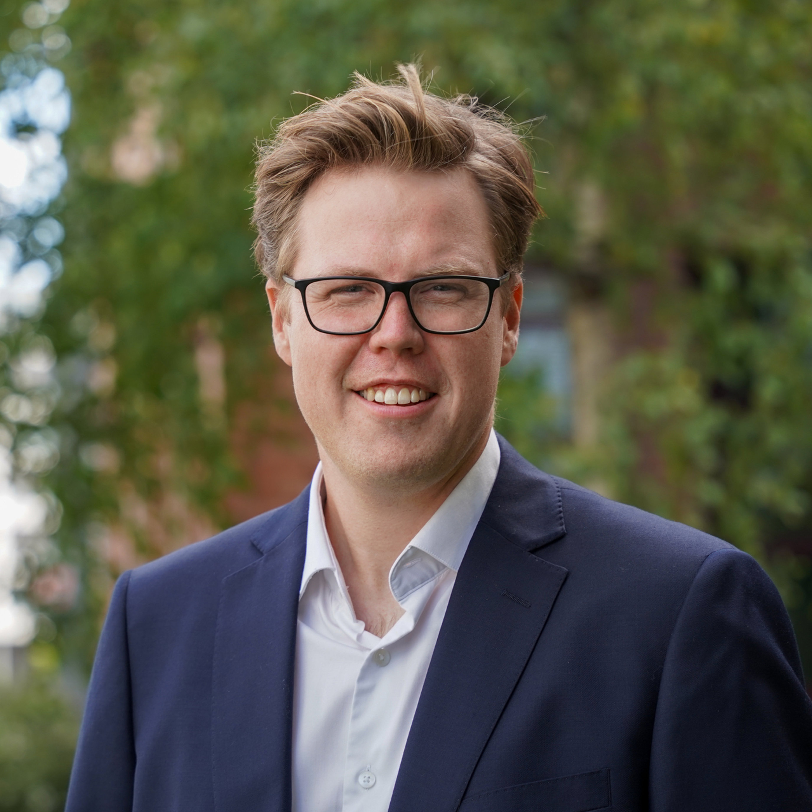 Mann med briller i dress smiler utandørs med grønt lauvverk i bakgrunnen.