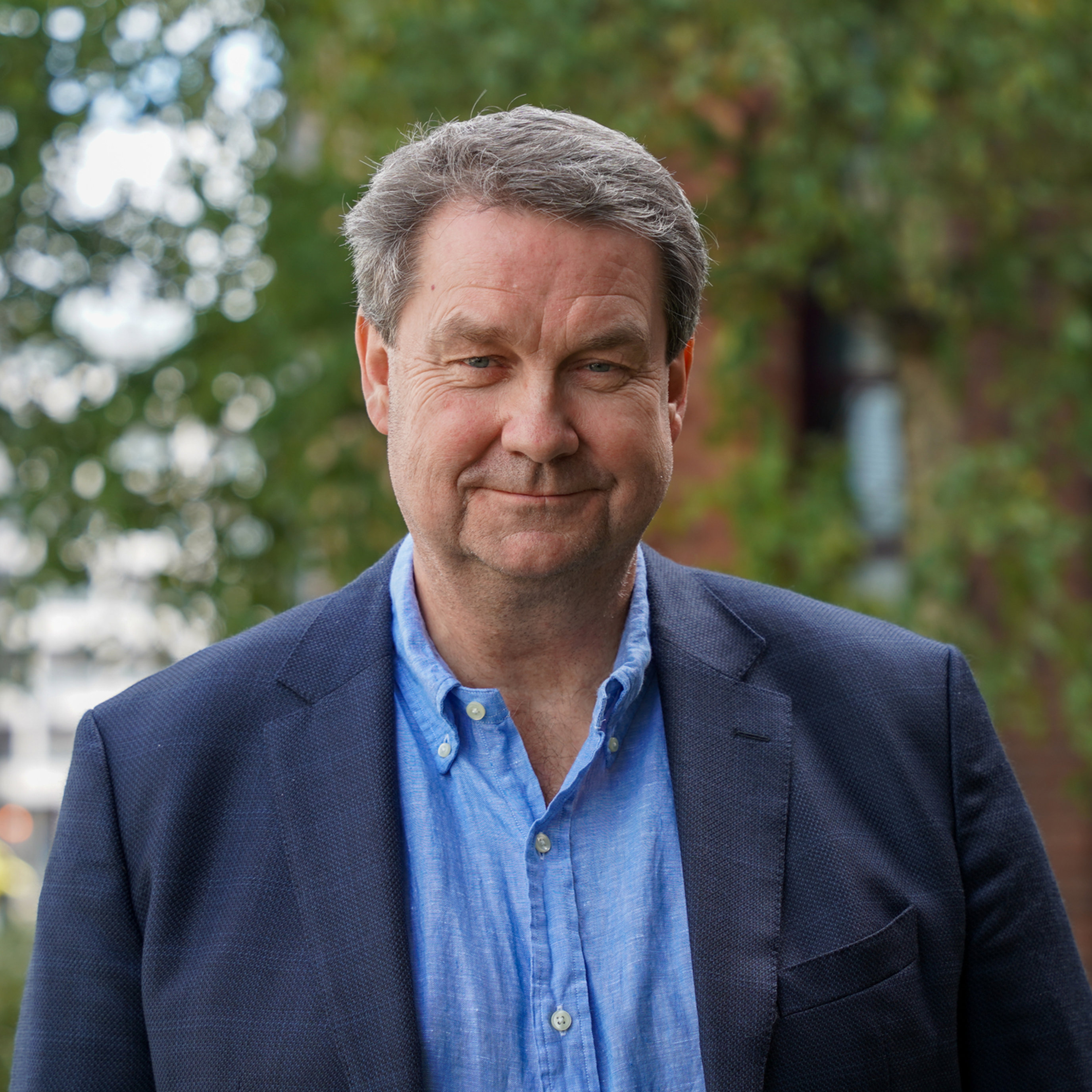 Ein mann i blå skjorte og blazer står utandørs framfor grøne tre.