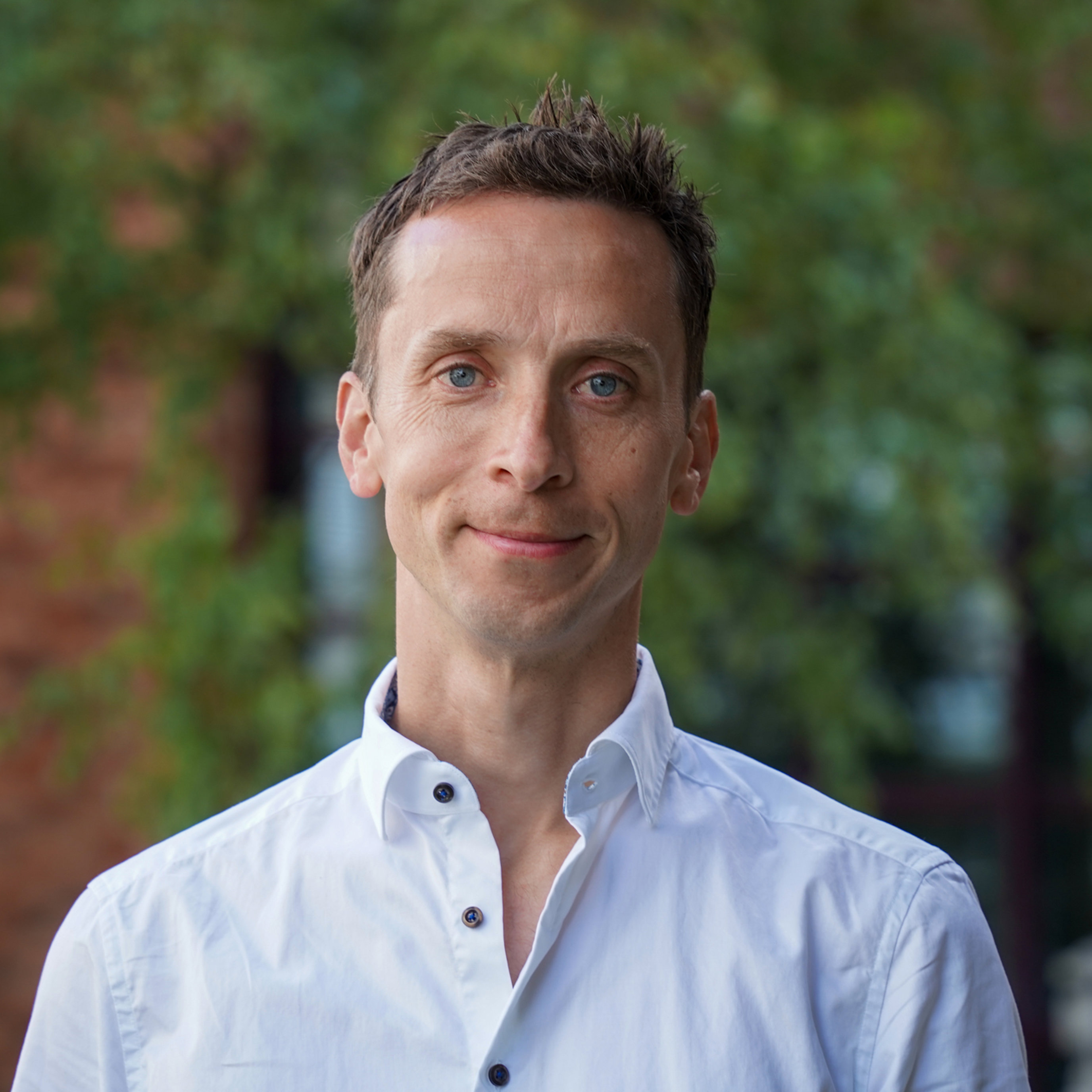 Mann i kvit skjorte med bakgrunn av grønt lauv og murbygning.