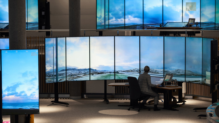 Person som sit foran fleire store skjermer på eit kontrollsenter, med bilete av snødekte fjellandskap som blir viste på skjermane.