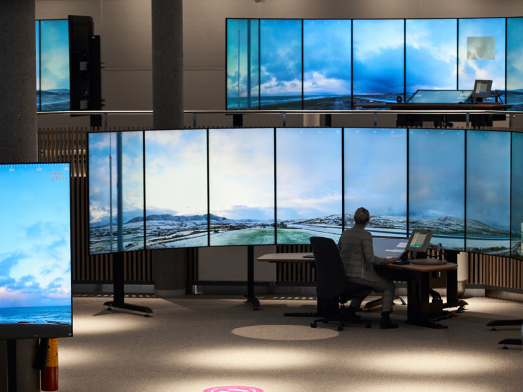 Kontrollrom med mange skjermer som viser landskapsbilder, hvor en person sitter foran en stor skjermvegg.