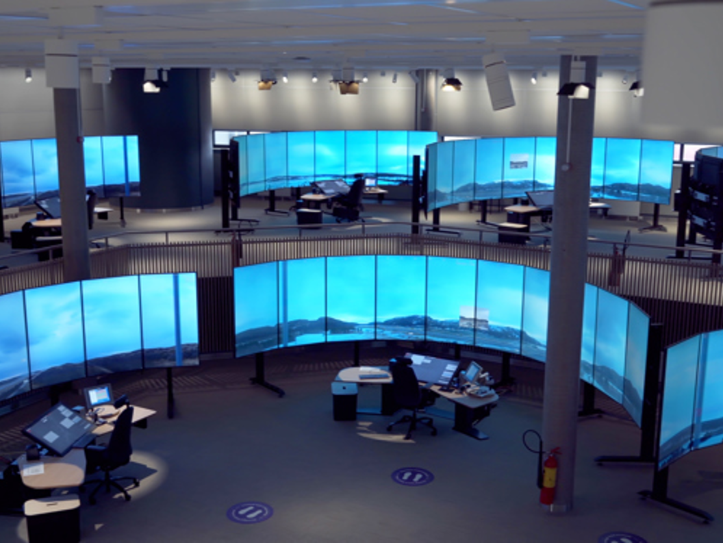 Kontrollrom med store, kurva skjermer som viser landskapsbilde. Fleire arbeidsstasjonar med kontorutstyr synest i rommet