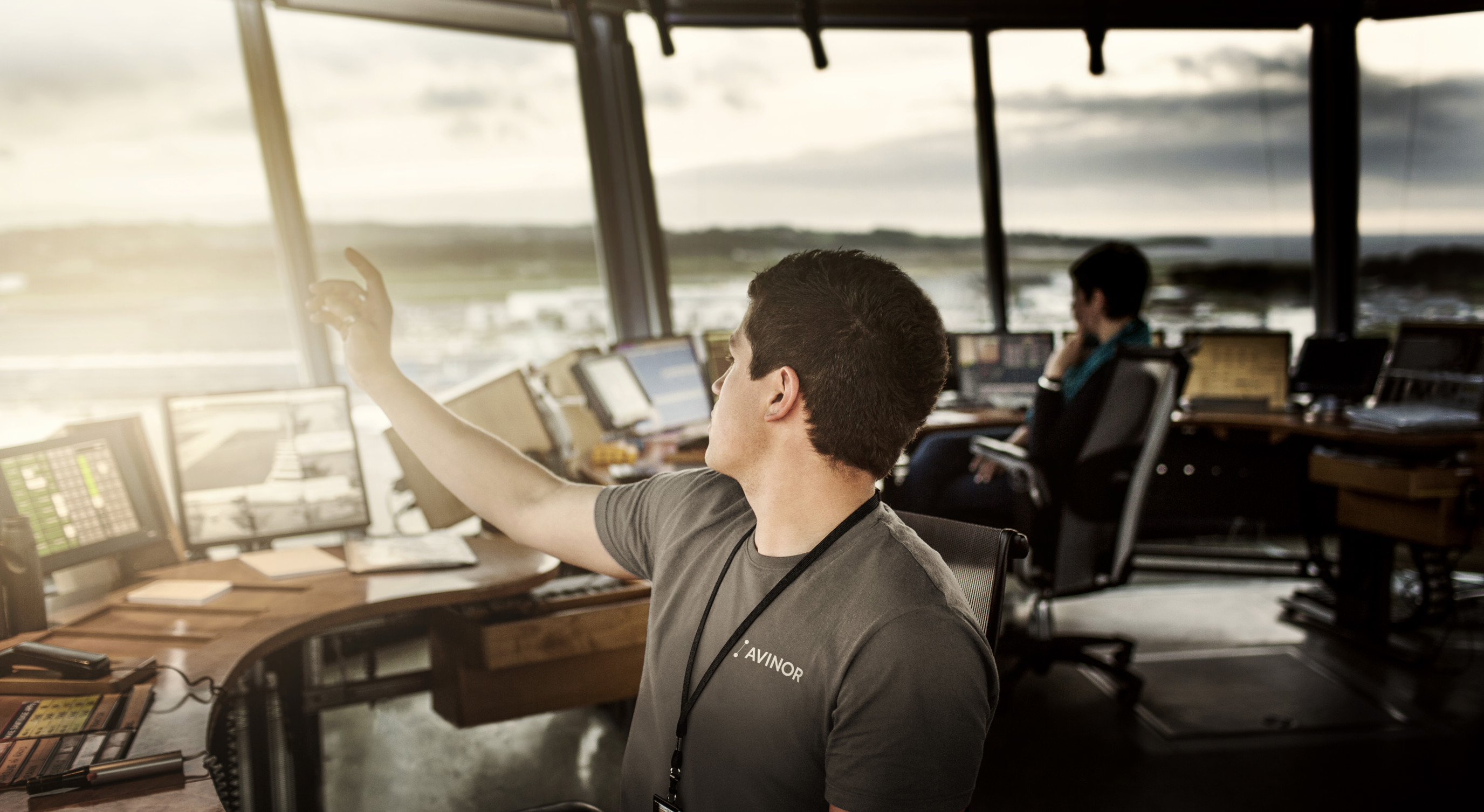 Mann i tårnkontroll med skjermer, peker ut av vinduet mens en kollega jobber i bakgrunnen på en flyplass, solnedgang i horisonten.