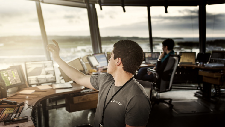 Mann i tårnkontroll med skjermar, peikar ut av vindauget medan ein kollega jobbar i bakgrunnen på ein flyplass, solnedgang i horisonten.