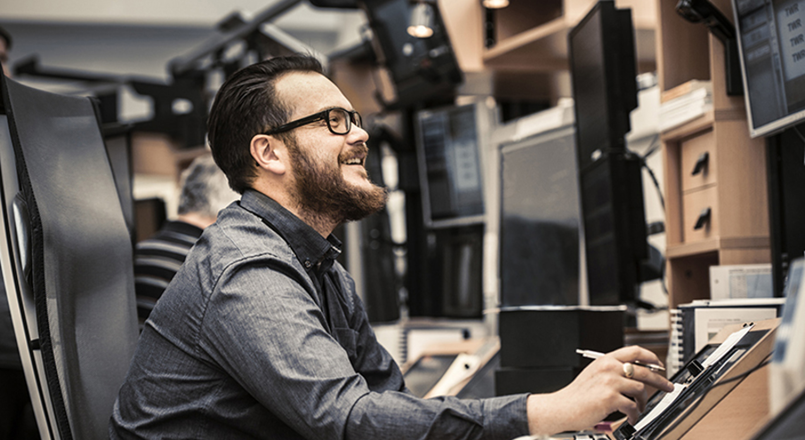 Mann med skjegg og briller smiler mens han arbeider på en datamaskin i et kontormiljø med flere skjermer.