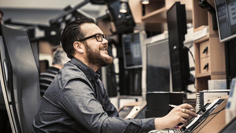 Mann med skjegg og briller smiler medan han arbeider på ein datamaskin i eit kontormiljø med fleire skjermar.