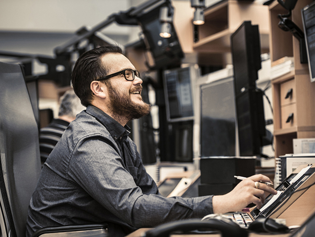 Smilende mann som jobber ved et skrivebord med flere dataskjermer, bruker en digital penn, i et moderne kontormiljø