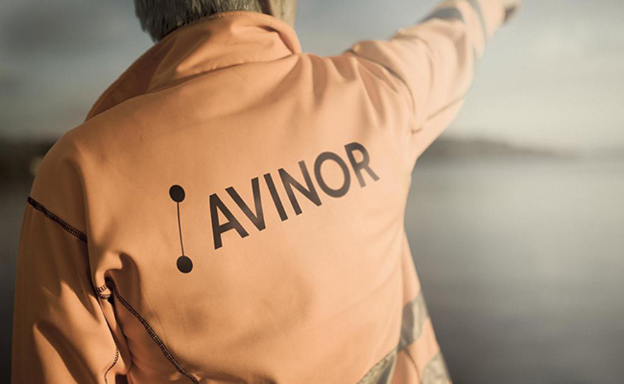 A man with an orange work jacket with the Avinor logo on the back pointing upwards towards the clouds, focus on the logo.