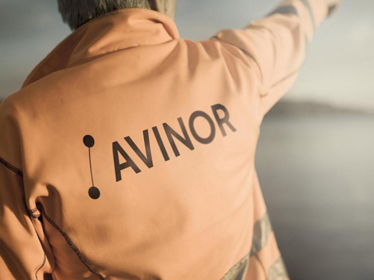 Ein mann med oransje arbeidsjakke med Avinor-logo på ryggen som peiker oppover mot skyene, fokus på logoen.