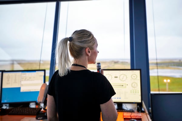 Air traffic controller in the tower with her back turned while communicating via radio, looking out the window towards the runway. Two computer screens display air traffic data.