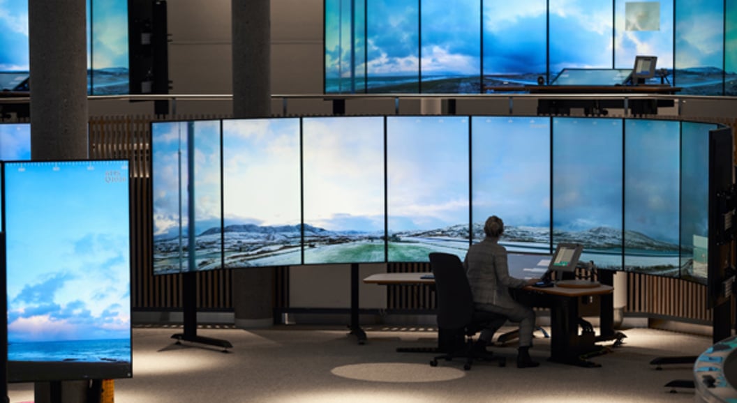 Ein person sit ved eit kontrollrom omgjeve av store skjermar som viser eit landskap av himmel og fjell.