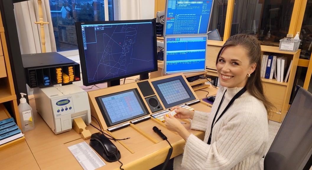 Smilende kvinne ved kontrollpult med dataskjermer i moderne operasjonsrom eller kontrollsenter, med teknisk utstyr og instrumentpaneler