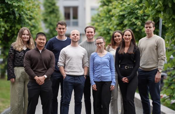 Ein gruppe unge vaksne står saman utandørs, omgitt av tre, alle kledde i casual klede. Dei ser mot kameraet og smiler